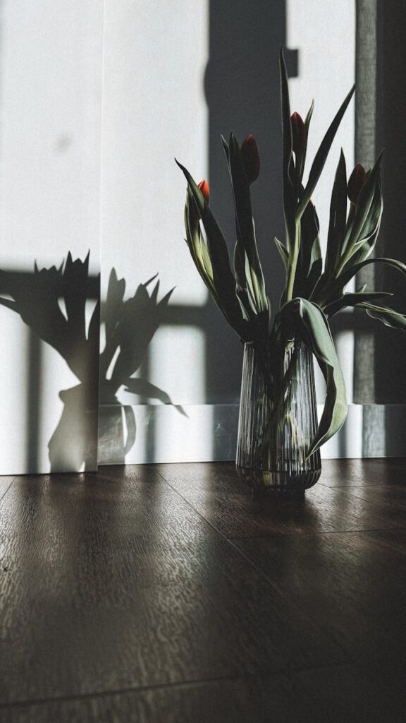 Aesthetic tulip arrangement in glass vase casting shadows on wooden floor.